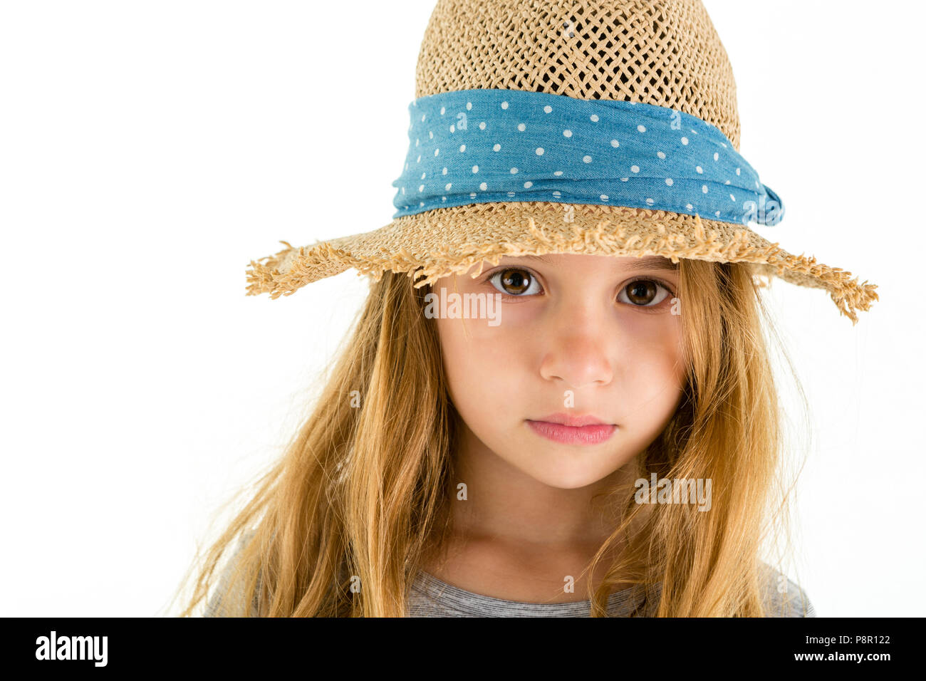 Adorable pretty little girl with big soulful eyes looking out from under the wide brim of a straw hat at the camera with a solemn expression, isolated Stock Photo