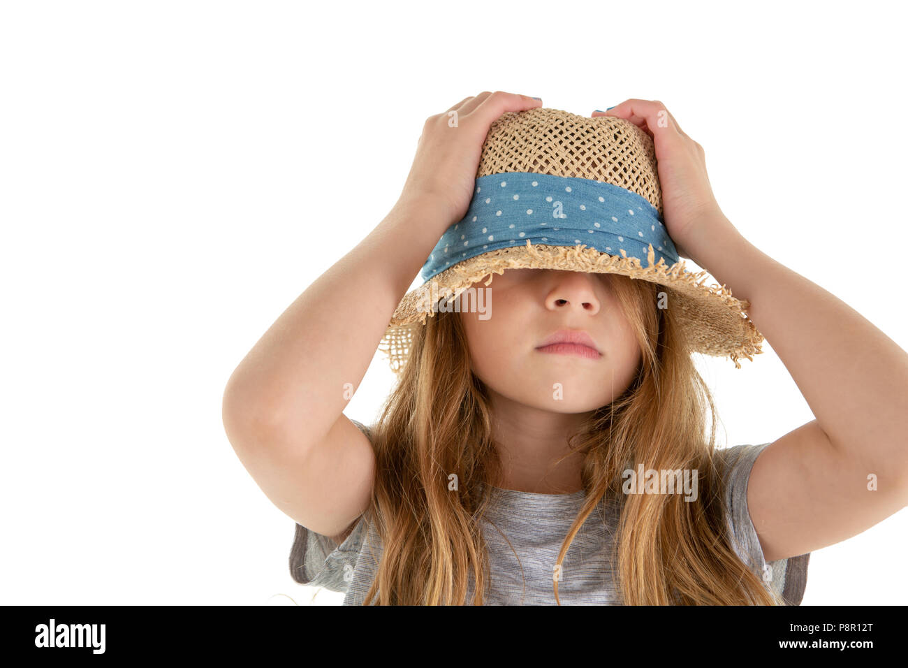 Funny little girl hiding underneath her straw hat pushing the crown down on her head with her hands in embarrassment isolated on white Stock Photo