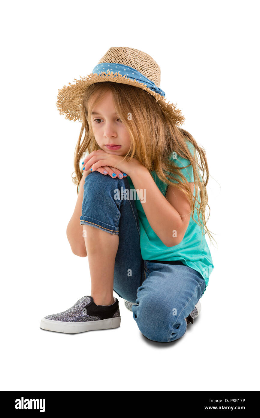 Melancholy little girl in a trendy straw hat and jeans kneeling staring at the ground with a sombre depressed expression isolated on white Stock Photo