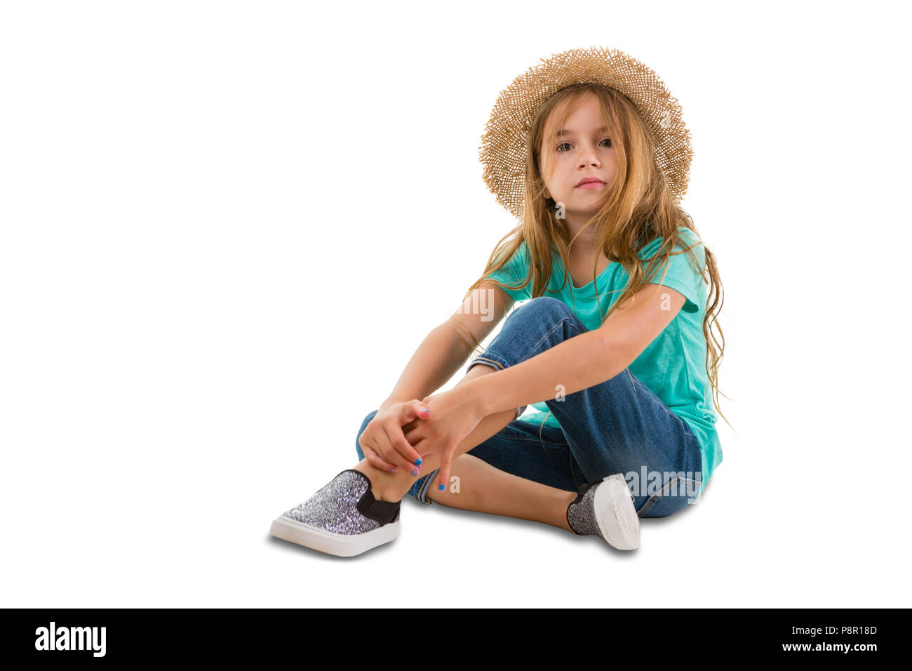 Serious little girl in trendy denims and a straw hat sitting relaxing on the floor looking to the side, isolated on white Stock Photo