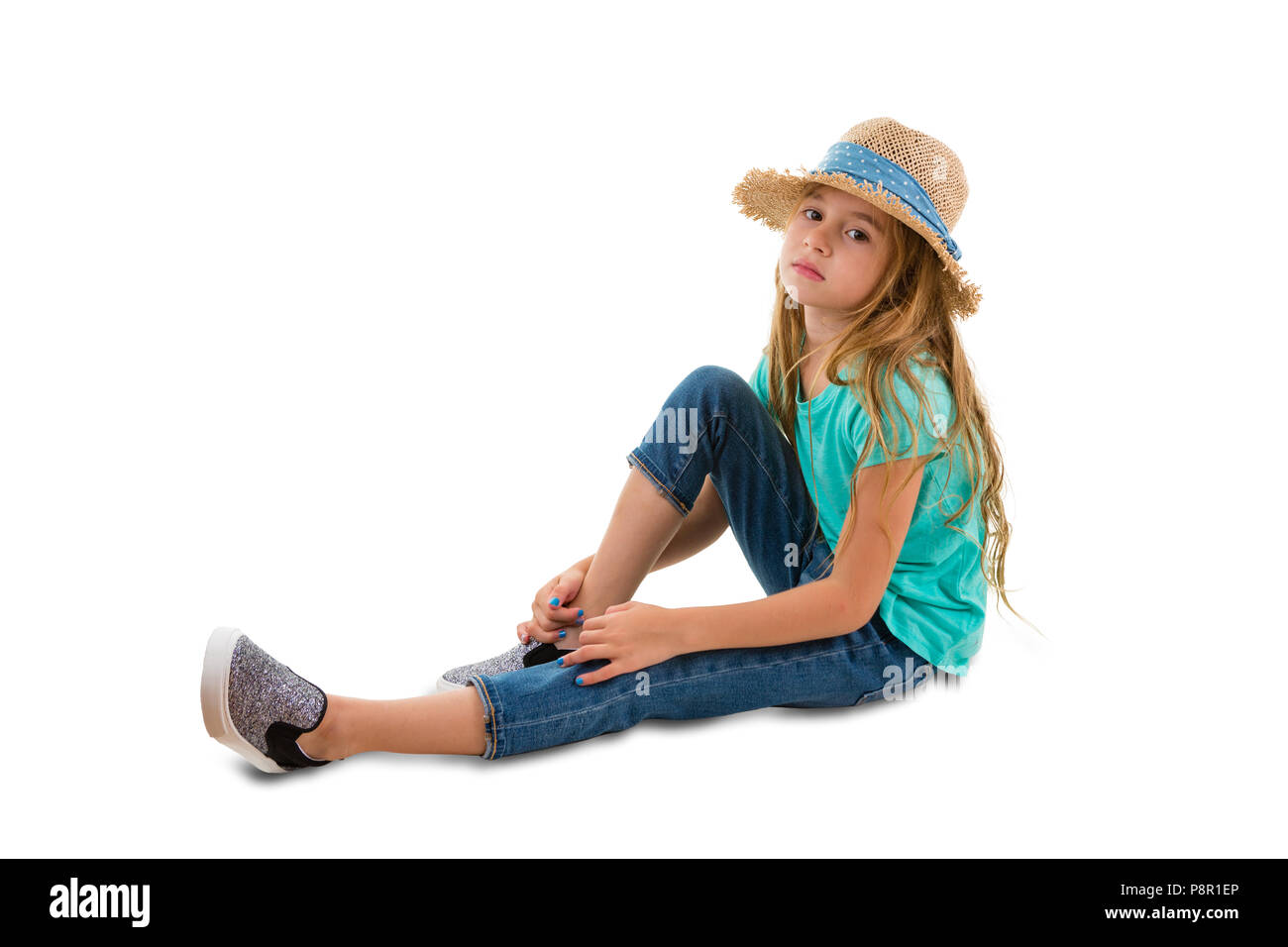 Solemn thoughtful little girl with long blond hair wearing a trendy straw hat sitting on the ground in a relaxed position watching the camera isolated Stock Photo