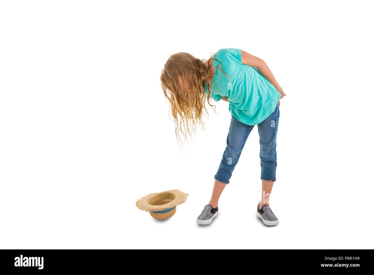 Little girl has dropped her straw hat on the ground bending over sideways to take a look with her long blond hair covering her face isolated on white. Stock Photo