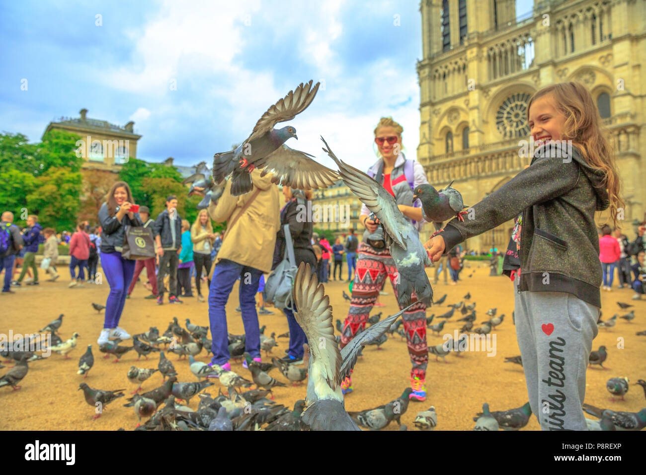 Paris, France - July 1, 2017: Happy child feeds pigeons in Notre Dame square full of people. Young tourist enjoys in Paris. Popular tourist destination in French capital. The Cathedral on background. Stock Photo
