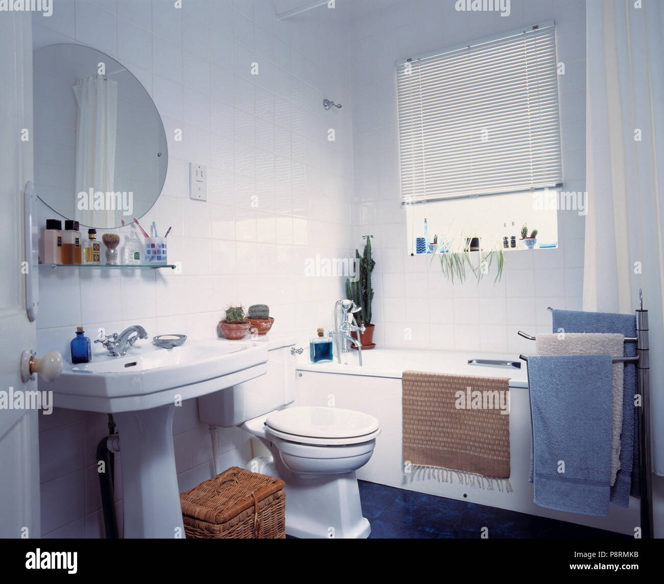 Circular mirror above pedestal basin in white tiled bathroom with blind above bath