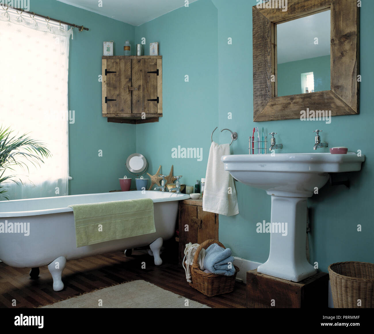 Rustic wooden mirror and corner cupboard in pale turquoise economy style bathroom with roll top bath and pedestal basin Stock Photo
