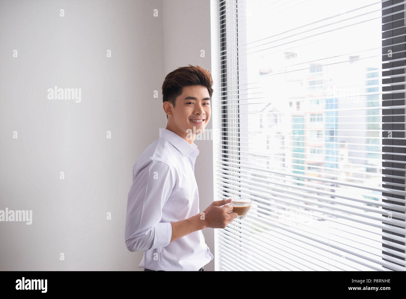 Portrait of a confident young businessman holding a cup of coffee while standing near office window, intelligent men in white shirt while resting afte Stock Photo