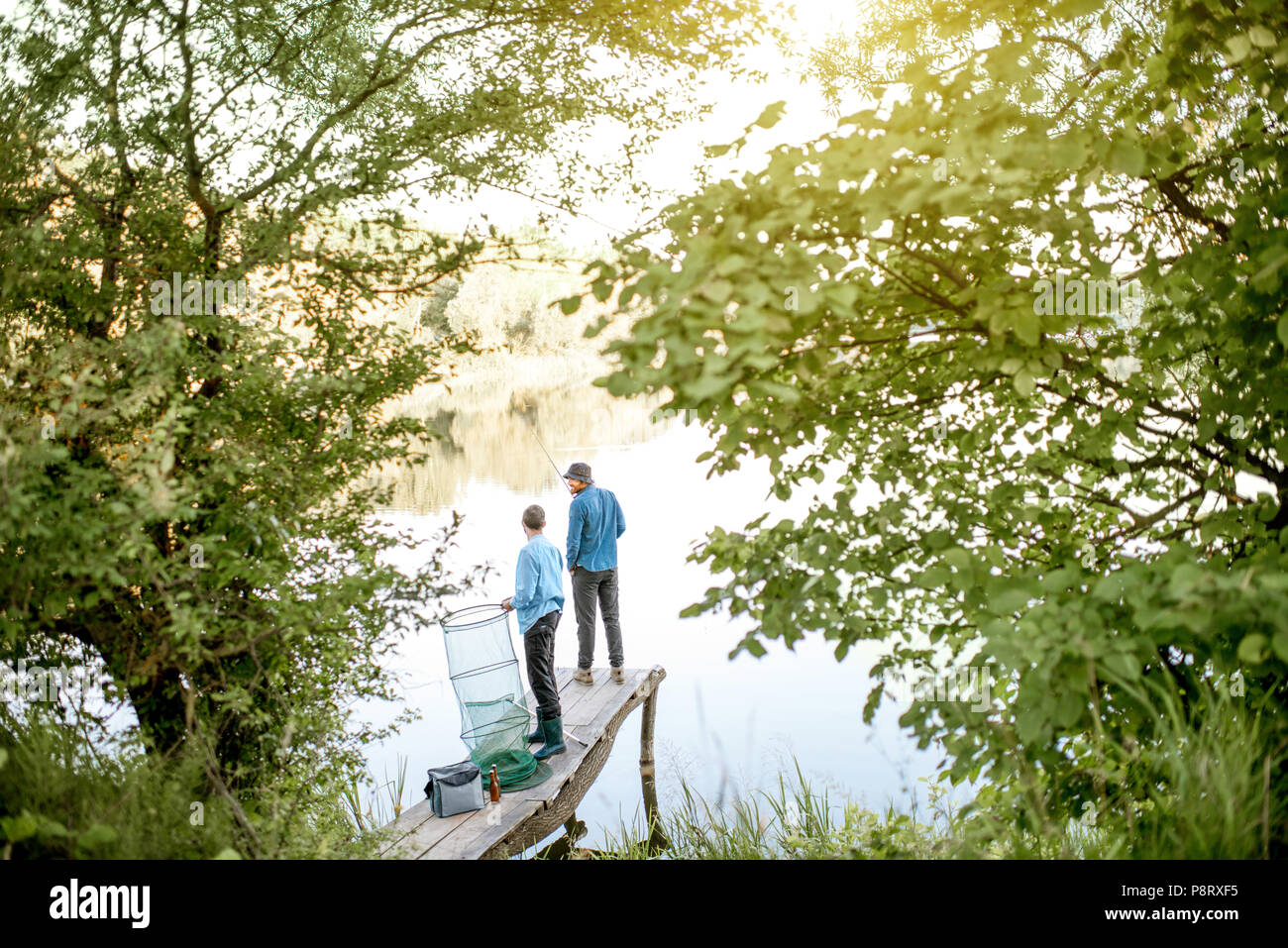 Two male friends preparing for fishing standing with fishing net and rod on the wooden pier during the morning light on the lake Stock Photo
