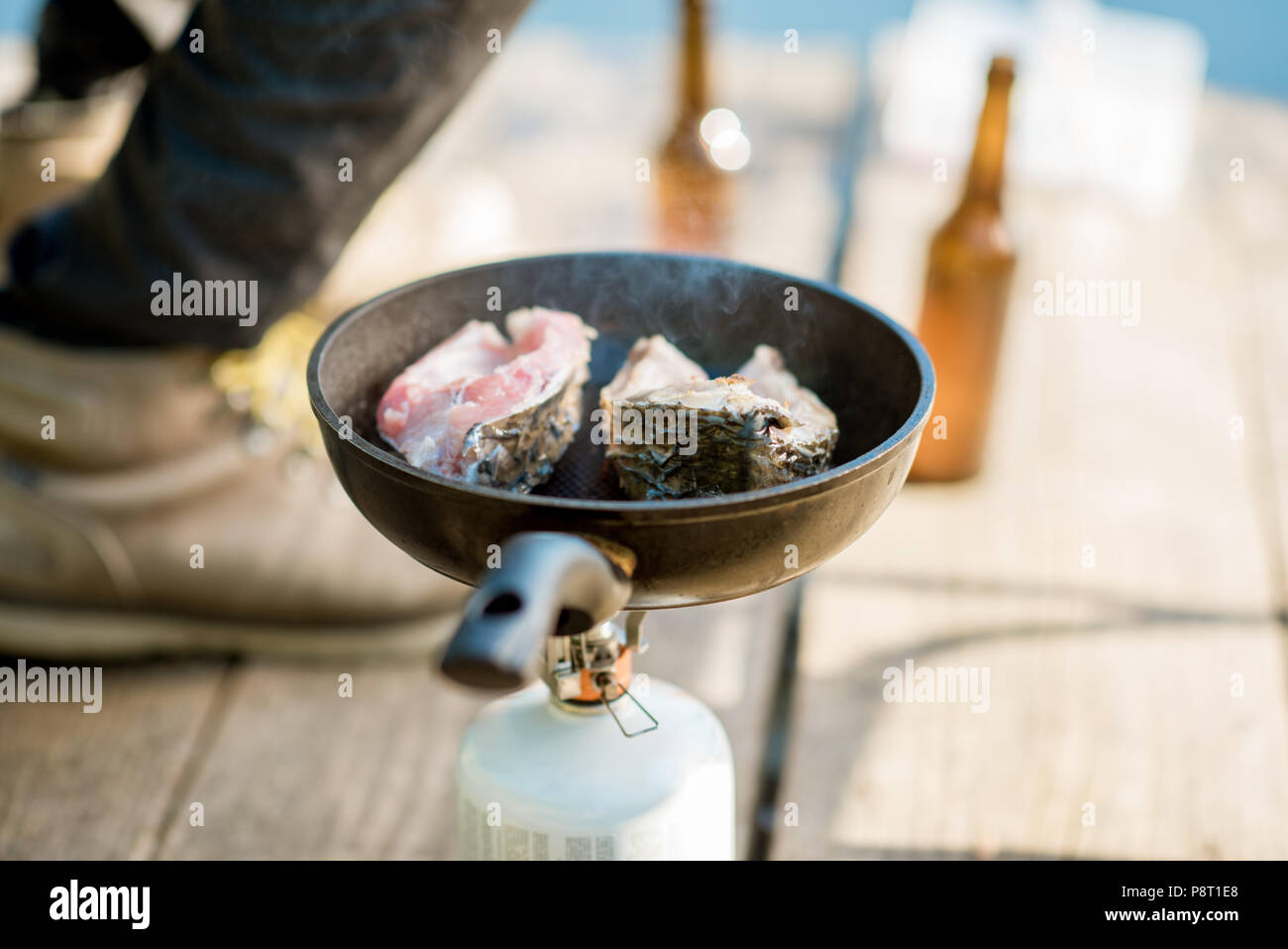 Frying two fish steakes on the burner during the picnic with beer and fishing tackles outdoors Stock Photo