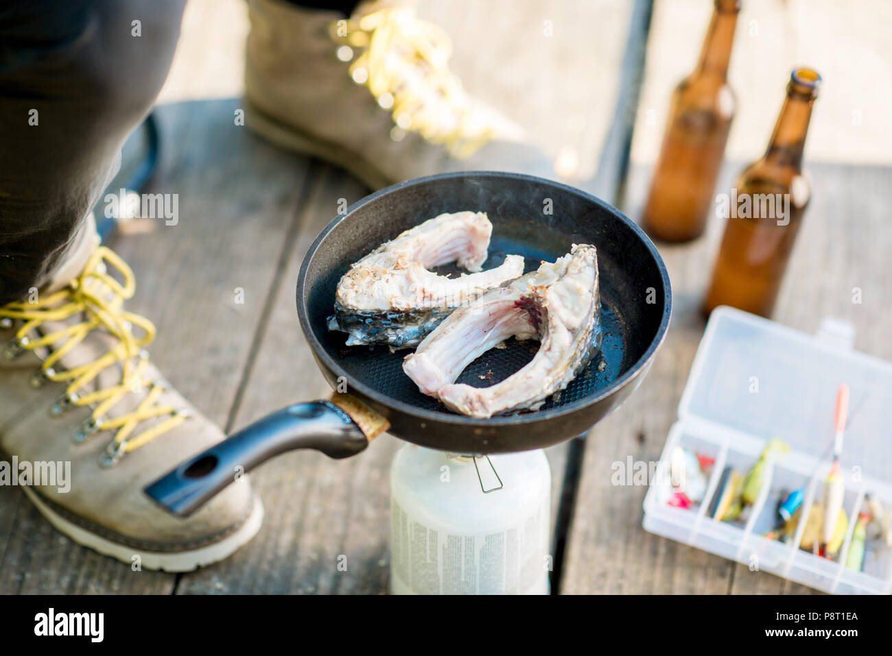 Frying two fish steakes on the burner during the picnic with beer and fishing tackles outdoors Stock Photo