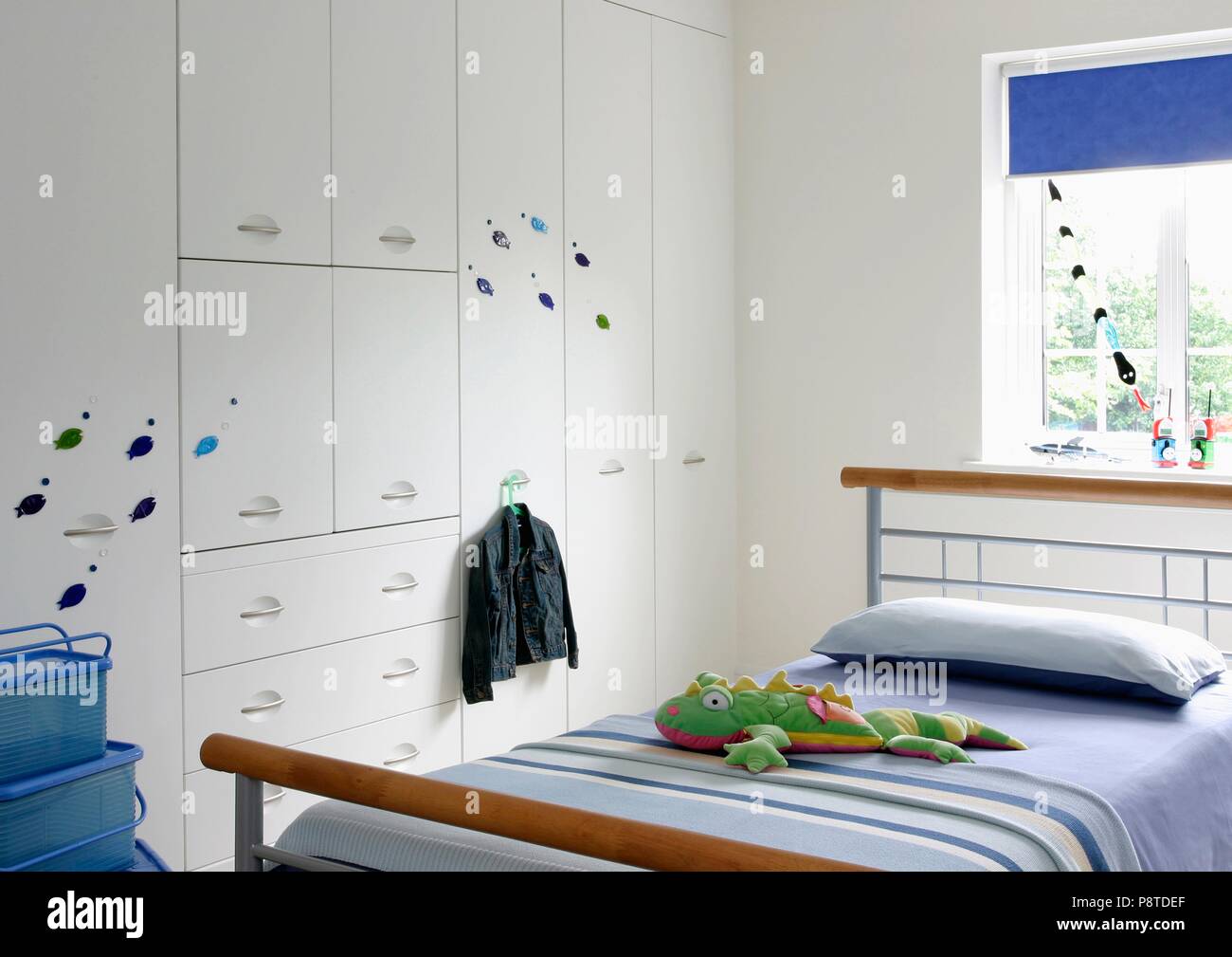 Blue striped cover on wood and metal bed in child's modern white bedroom with white fitted storage cupboard Stock Photo