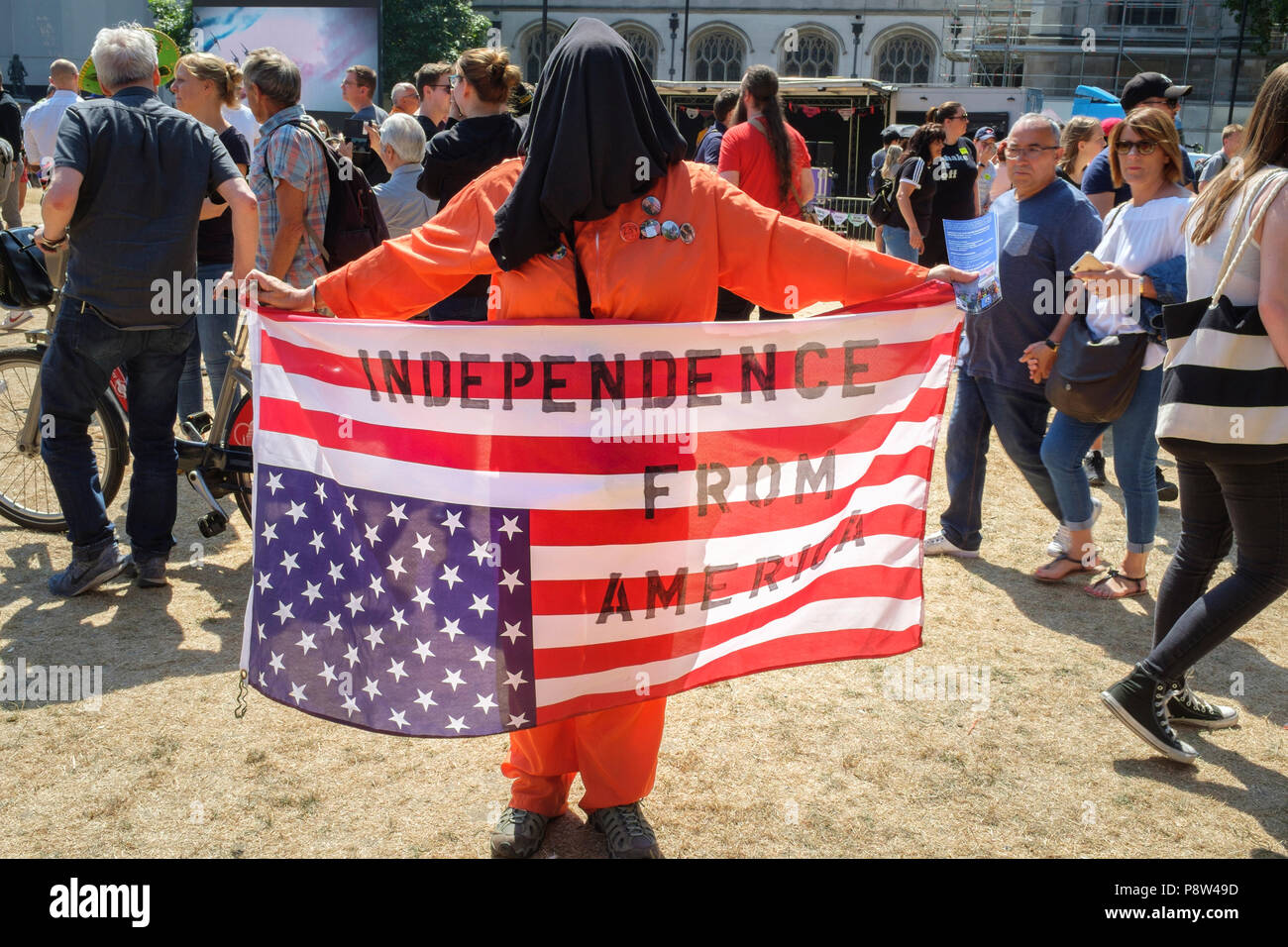 London, UK. 13th July 2018. Tens of thousands of people took to the streets of central London to protest against US President Donald Trump's visit to the UK. Pictured: A hooded and orange jumpsuit clad figure holds an inverted US flag with the words Independence from America stencilled on it. Credit: mark phillips/Alamy Live News Stock Photo