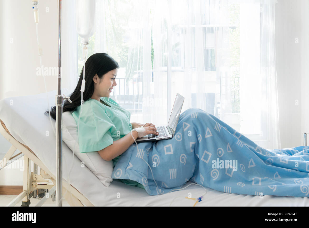Asian woman sick patient lying in a hospital bed using a laptop for relax when she recovering for ill in hospital. Stock Photo