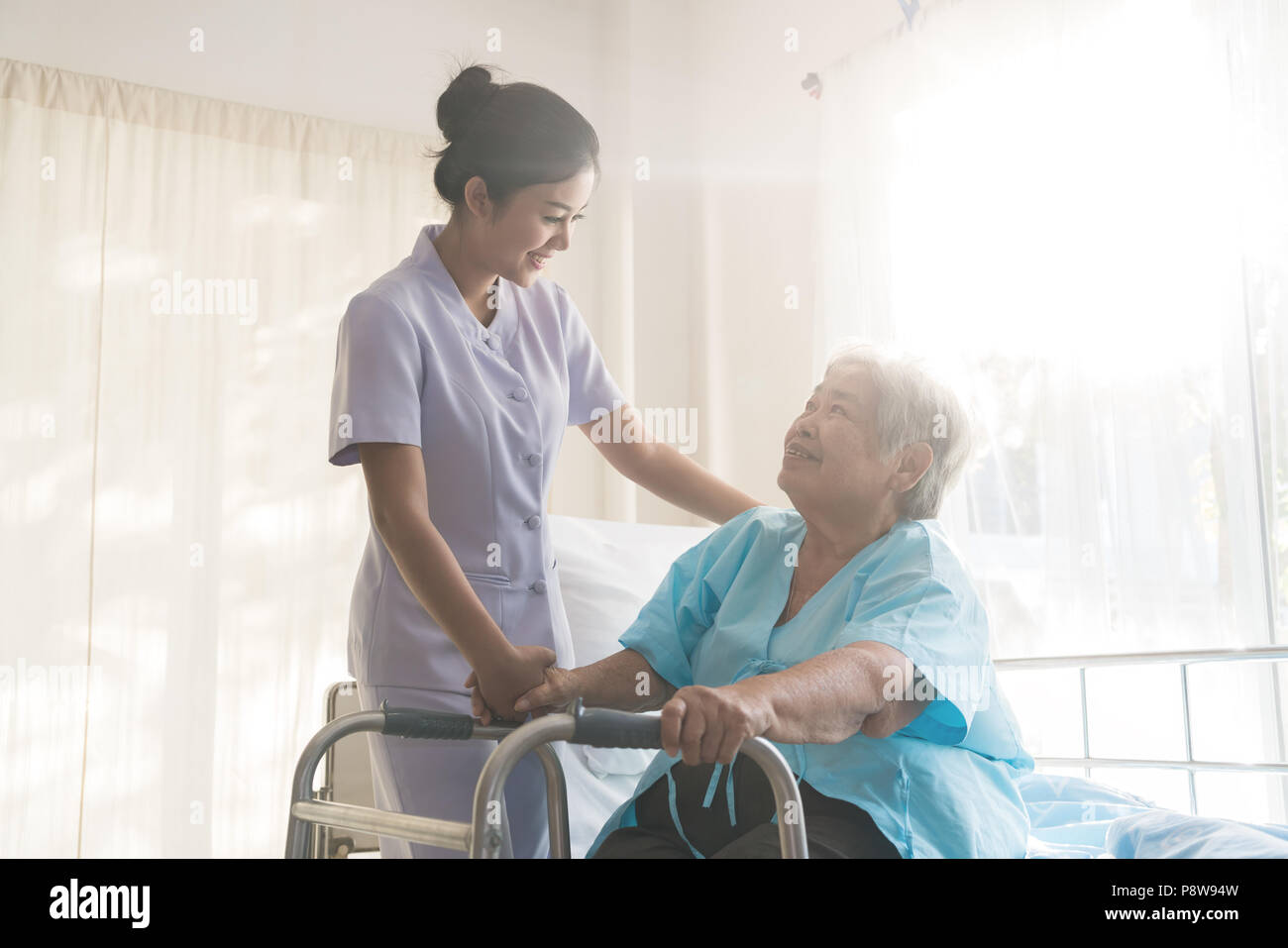 Asian young nurse supporting elderly patient disabled woman in using walker in hospital. Elderly patient care concept. Stock Photo
