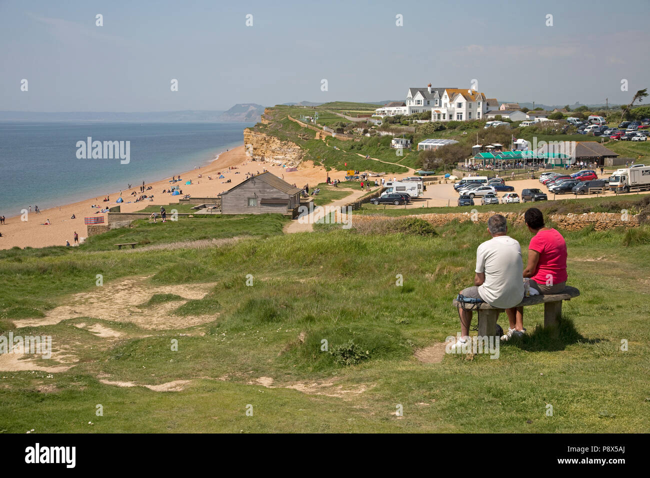 Couple sitting on seat overlooking Holiday makers on Hive Beach Burton Bradstock Dorset UK Stock Photo
