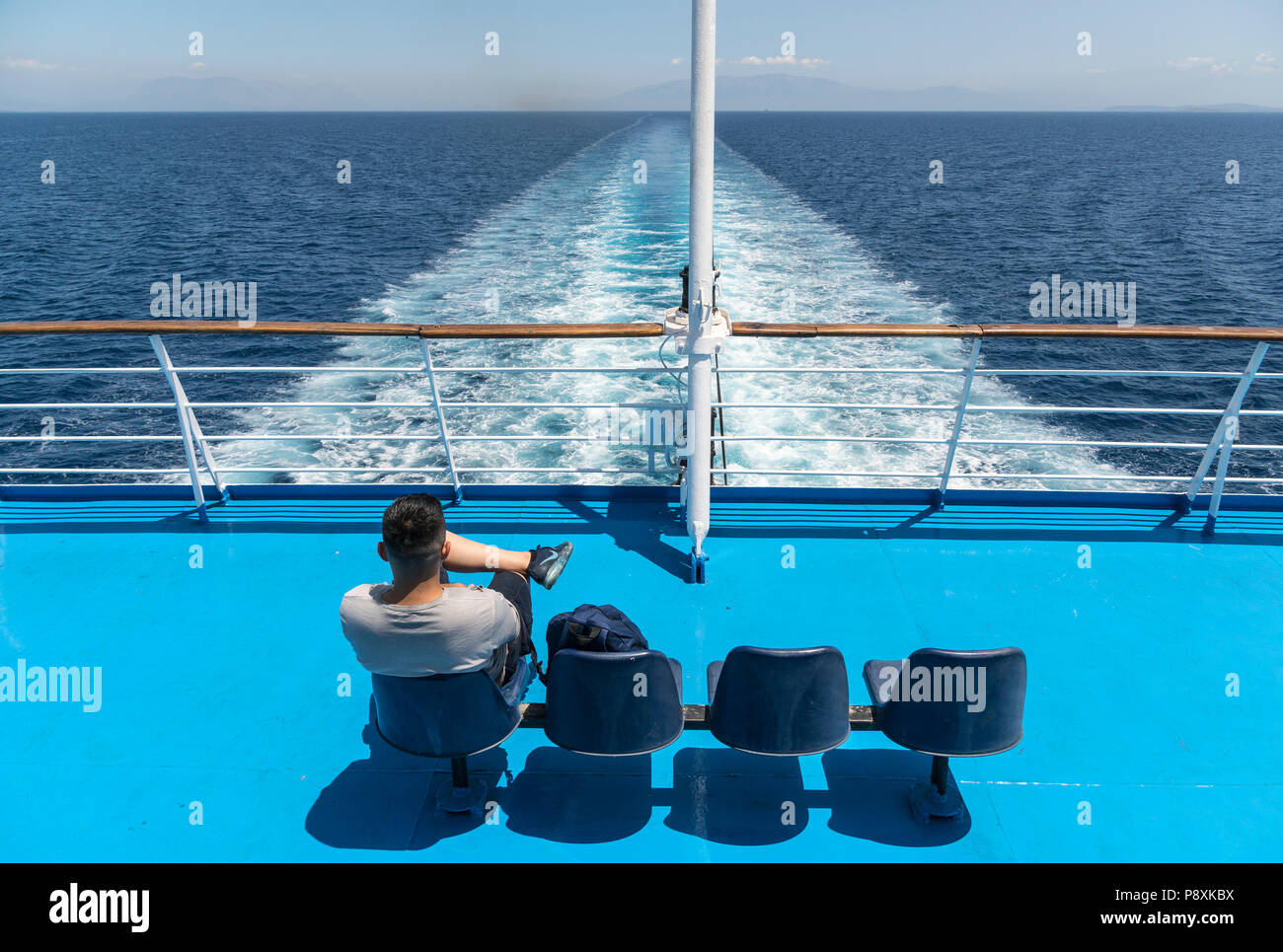 passenger on the stern of a Greek ferry watching the wake of the ship and the receding land. Stock Photo