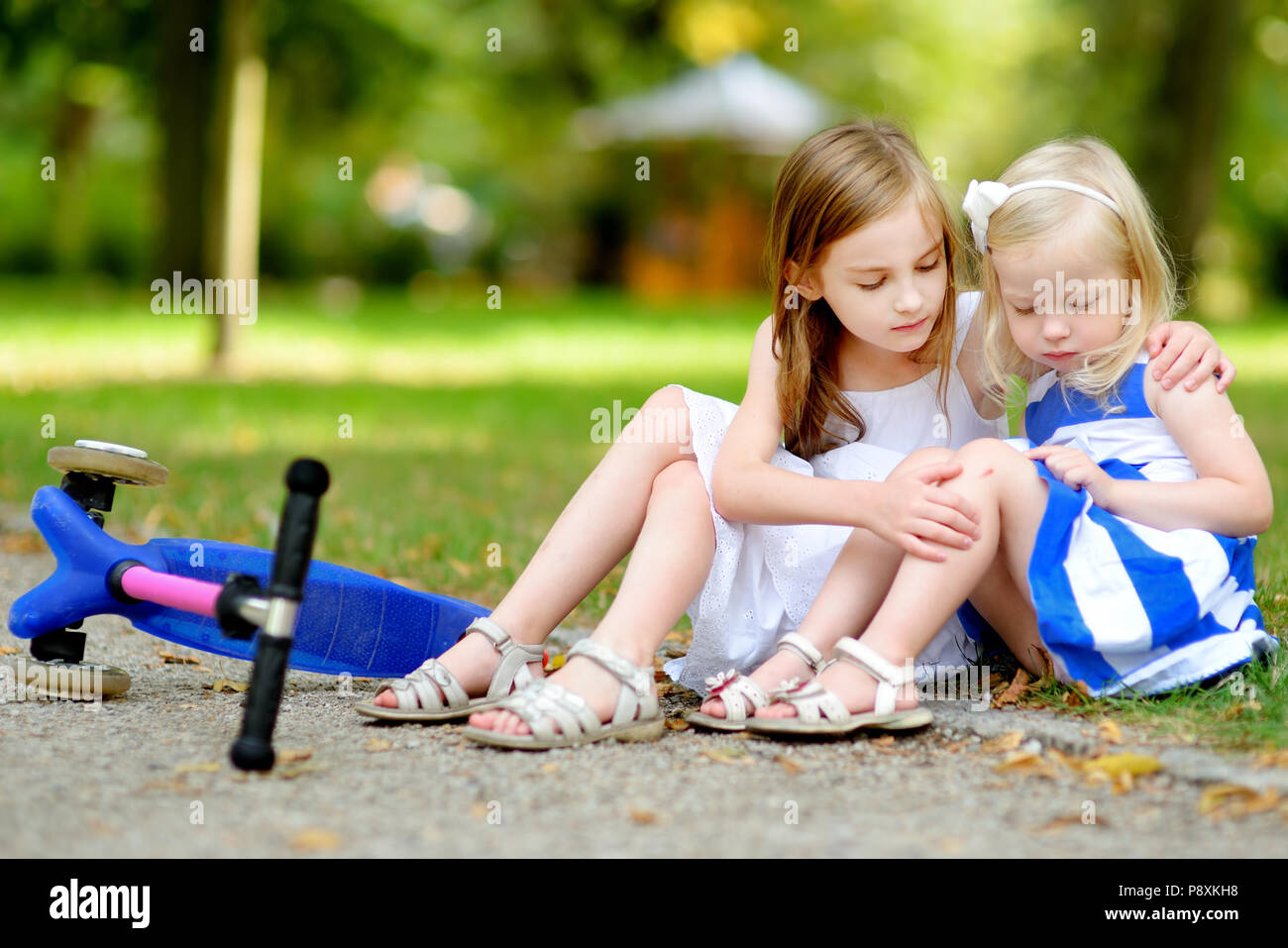 Little girl comforting her sister after she fell while riding her scooter at summer park Stock Photo