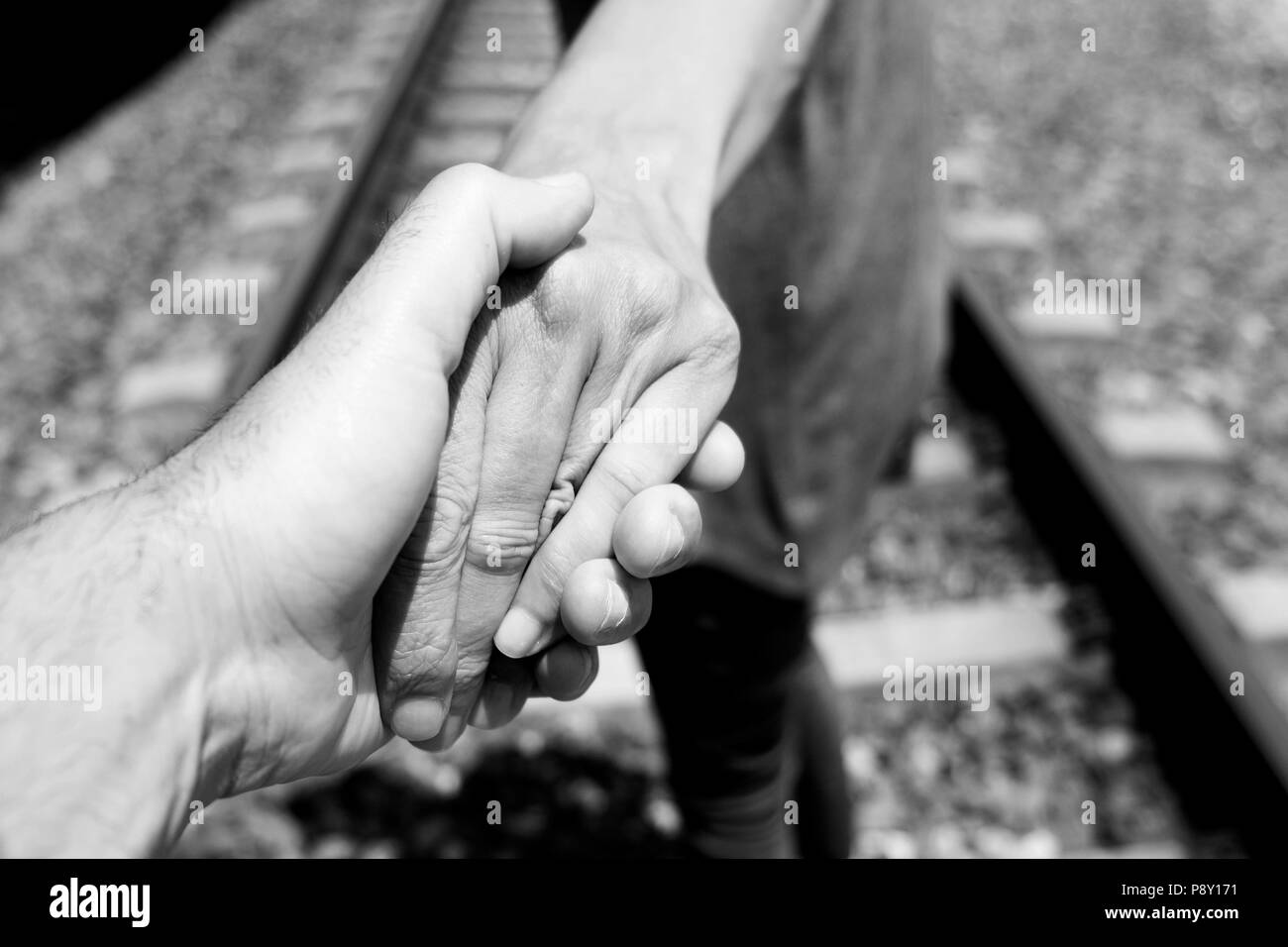 closeup of the hand of a caucasian man holding the hand of another caucasian man, seen from behind, walking by the railroad tracks, in black and white Stock Photo