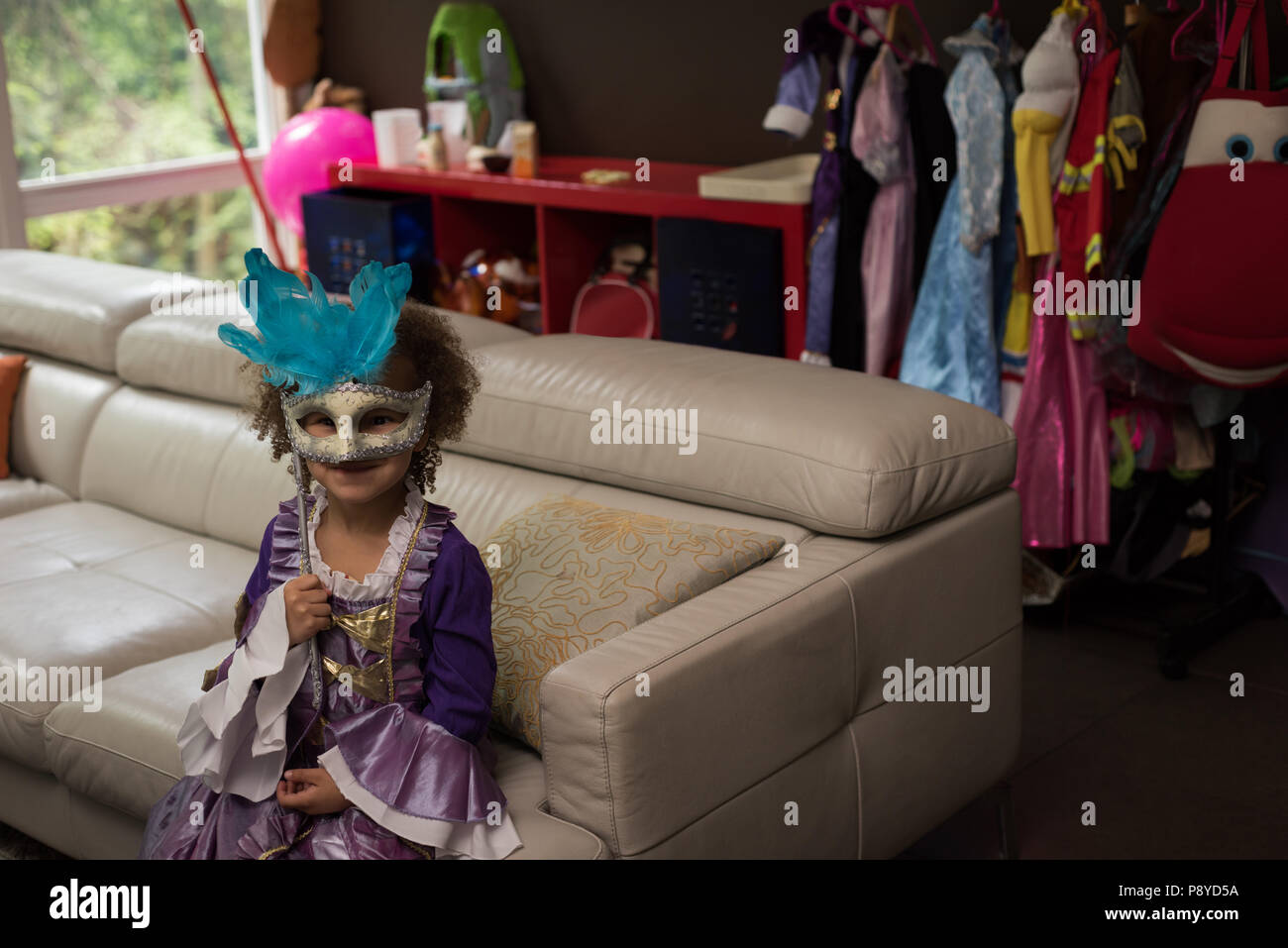 Girl wearing masquerade mask sitting on sofa Stock Photo