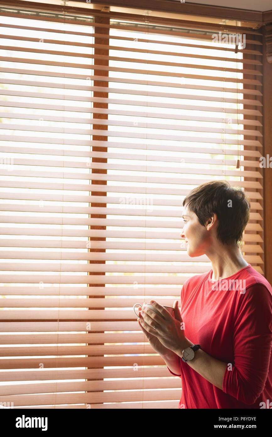 Woman looking through window while having coffee in living room Stock Photo