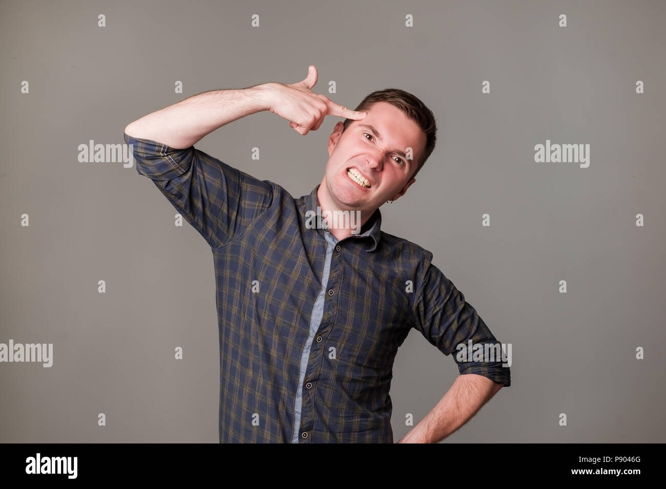 Tired or bored european guy holding gun gesture near temples and making faces while standing over gray background. Stock Photo