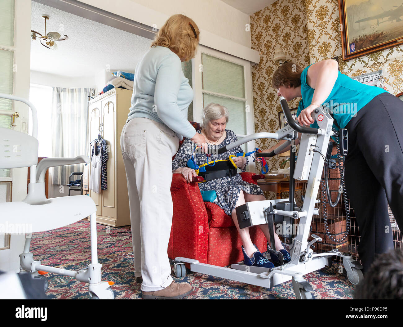 Old lady with dementia being hoisted from her recliner chair by two lady carers Stock Photo