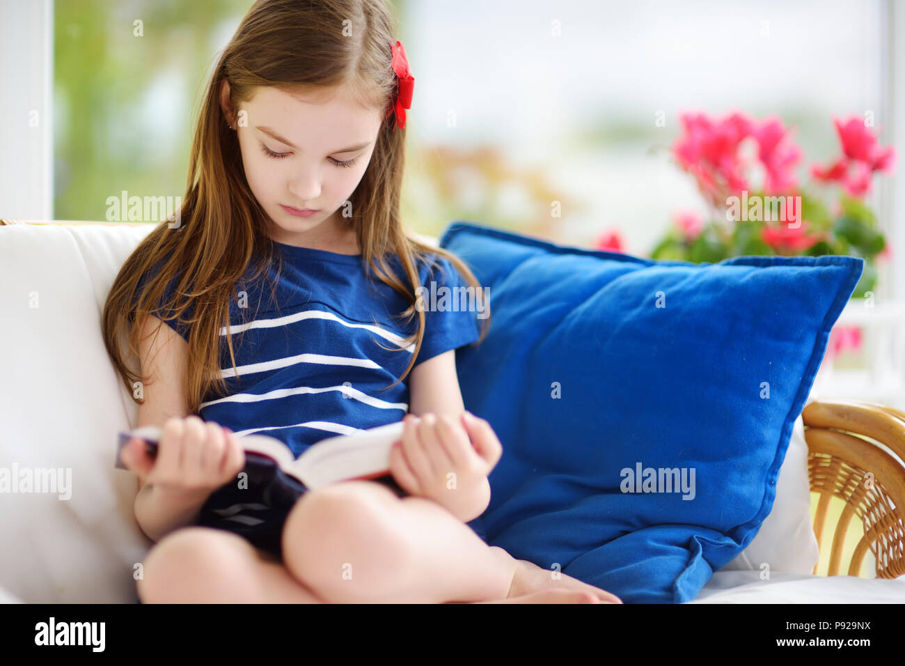 Adorable little girl reading a book in white living room on beautiful summer day. Smart schoolgirl doing her homework at home after school. Stock Photo