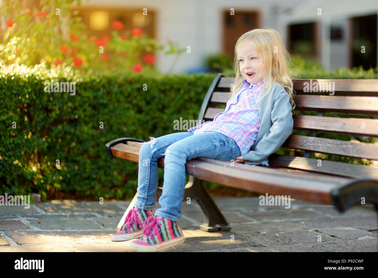 Cute little girl sitting on wooden bench in a city park on sunny summer ...