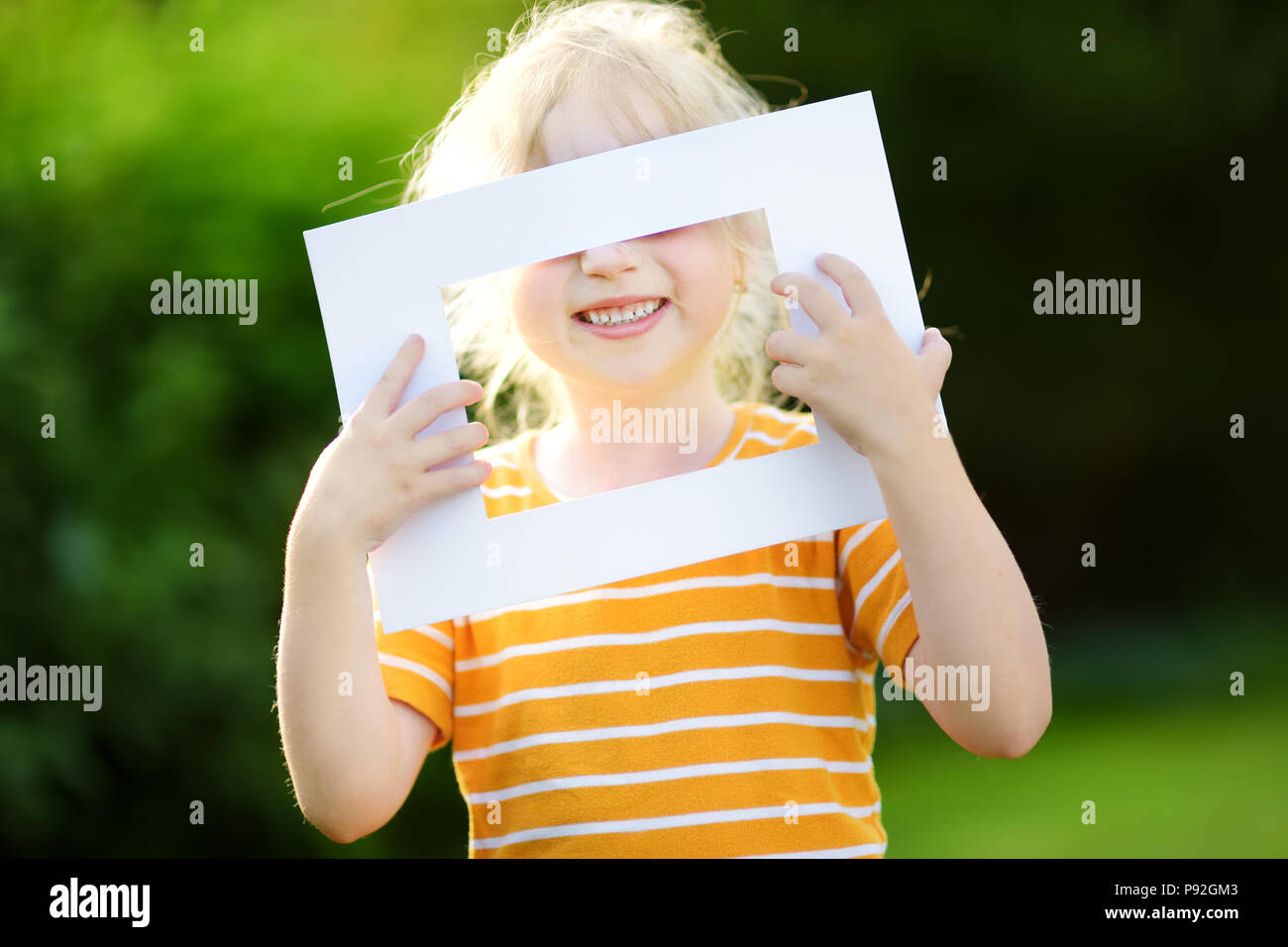 Cute cheerful little girl holding white picture frame in front of her face. Adorable child framing her smiling face. Stock Photo