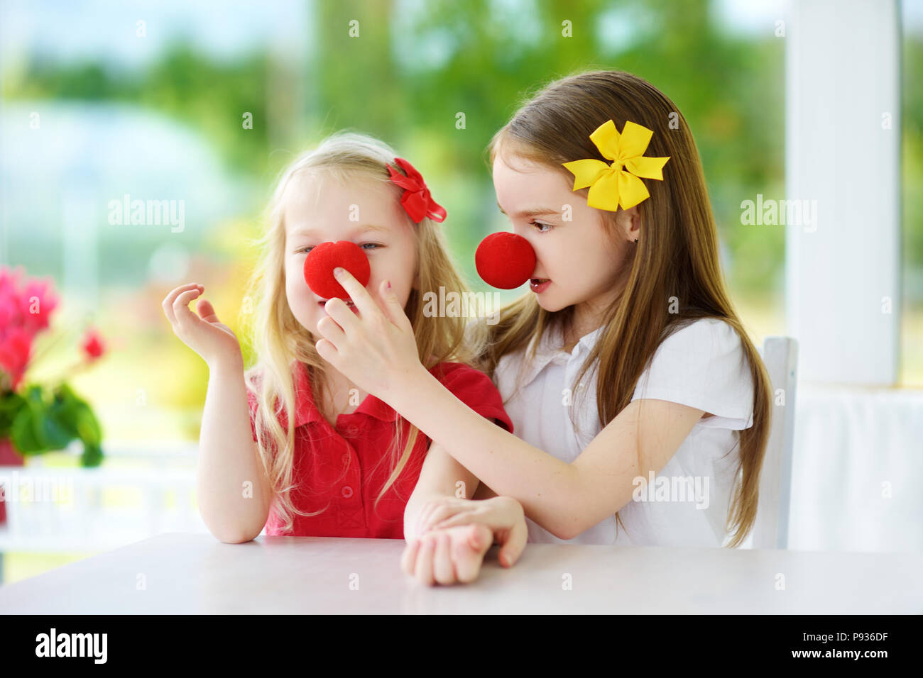 Happy little sisters wearing red clown noses having fun together on sunny summer day at home. Two kids playing together. Stock Photo