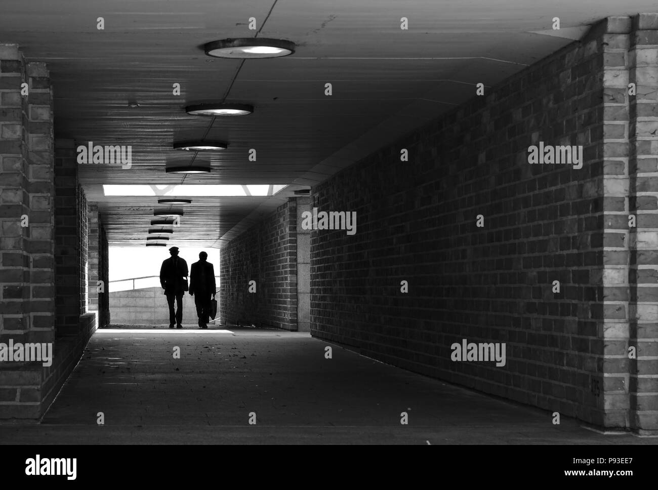 Two people silhouetted against a bright light walking through a tunnel with brick walls towards the camera (monochrome) Stock Photo