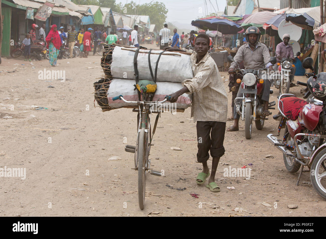 Kakuma, Kenya - Street scene. A boy transports charcoal rope cover on his bike on a busy dirt road. Stock Photo