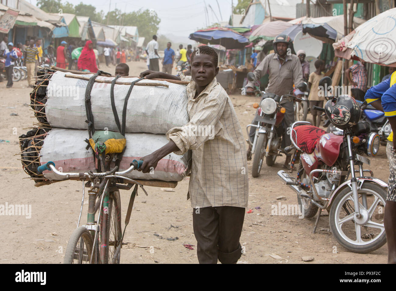 Kakuma, Kenya - Street scene. A boy transports charcoal rope cover on his bike on a busy dirt road. Stock Photo