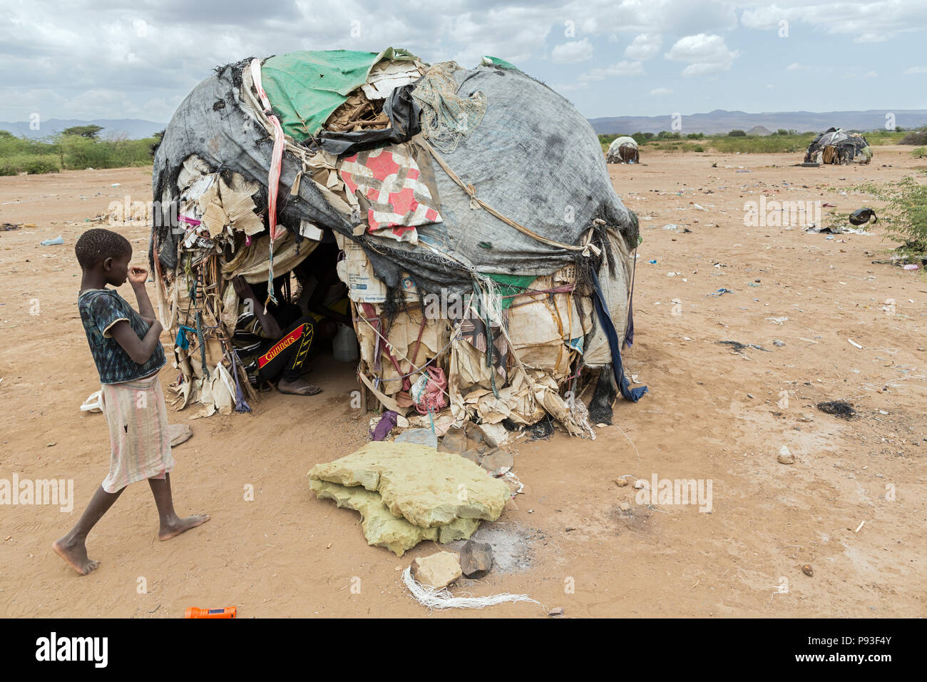 Kakuma, Kenya - On the edge of the refugee camp Kakuma. A girl is standing in front of a hut covered with old plastic tarpaulins, blankets, boxes and garbage. Stock Photo