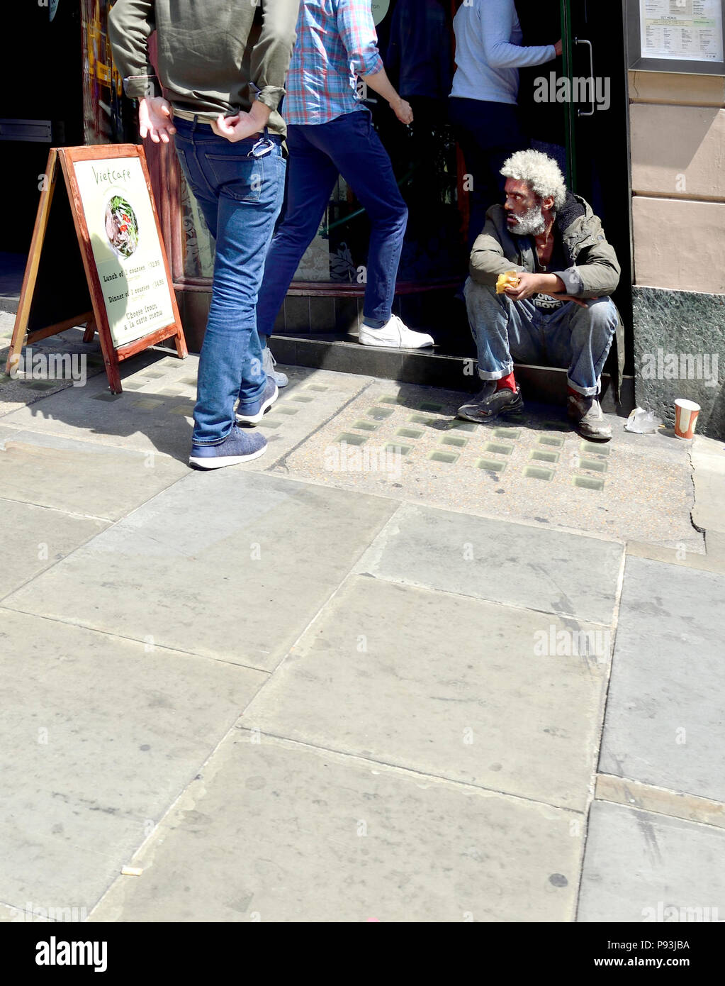 Black man with white hair and beard sitting on the step of a restaurant as people walk in past him, Whitehall, London, England, UK. Stock Photo