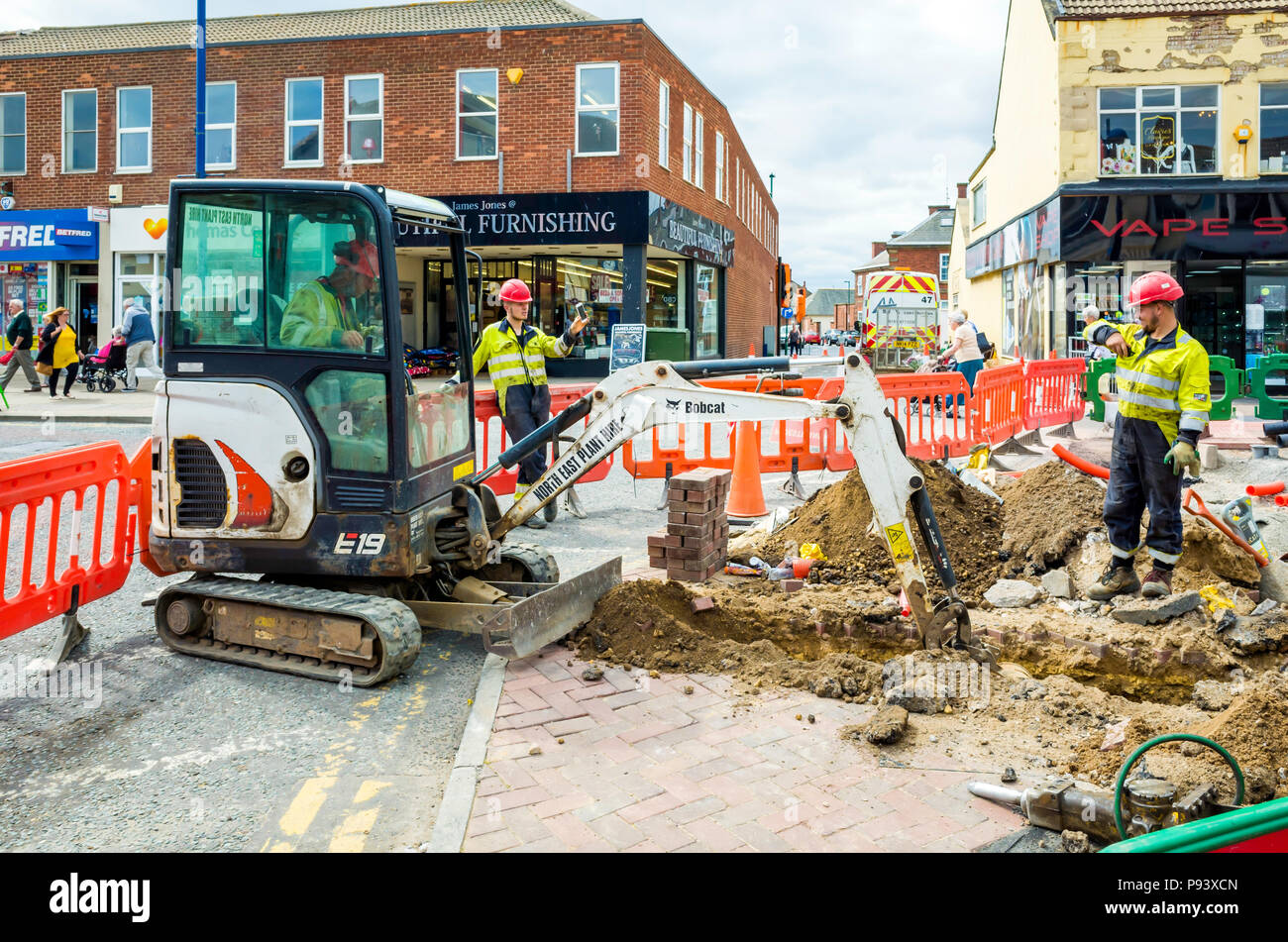 Workmen with a Bobcat excavator digging a trench in the pavement for installation of an electrical power cable Stock Photo
