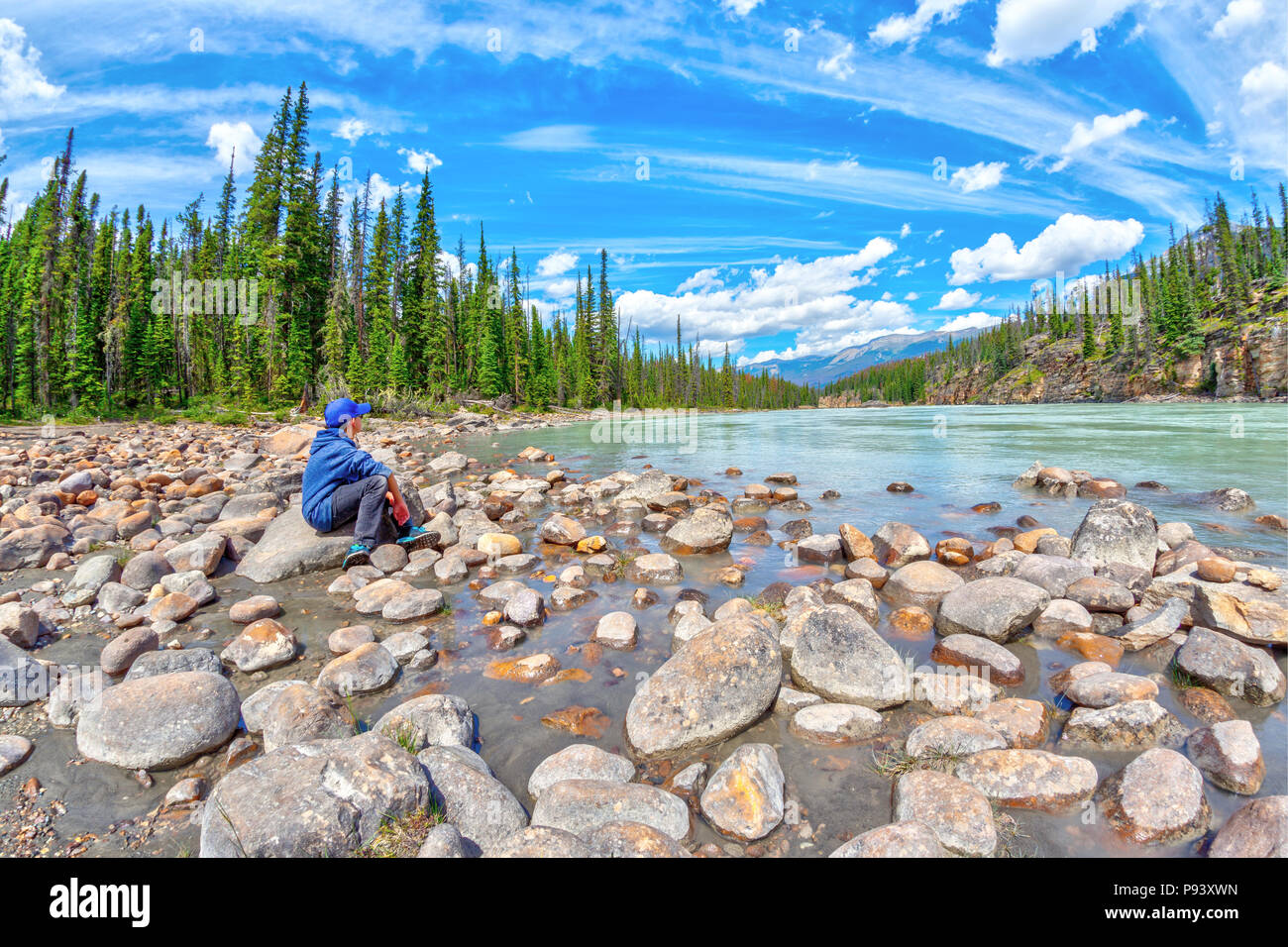 A boy sits on the edge of the Athabasca River resting and enjoying the tranquility in nature of the Canadian Rockies at Jasper National Park in Albert Stock Photo