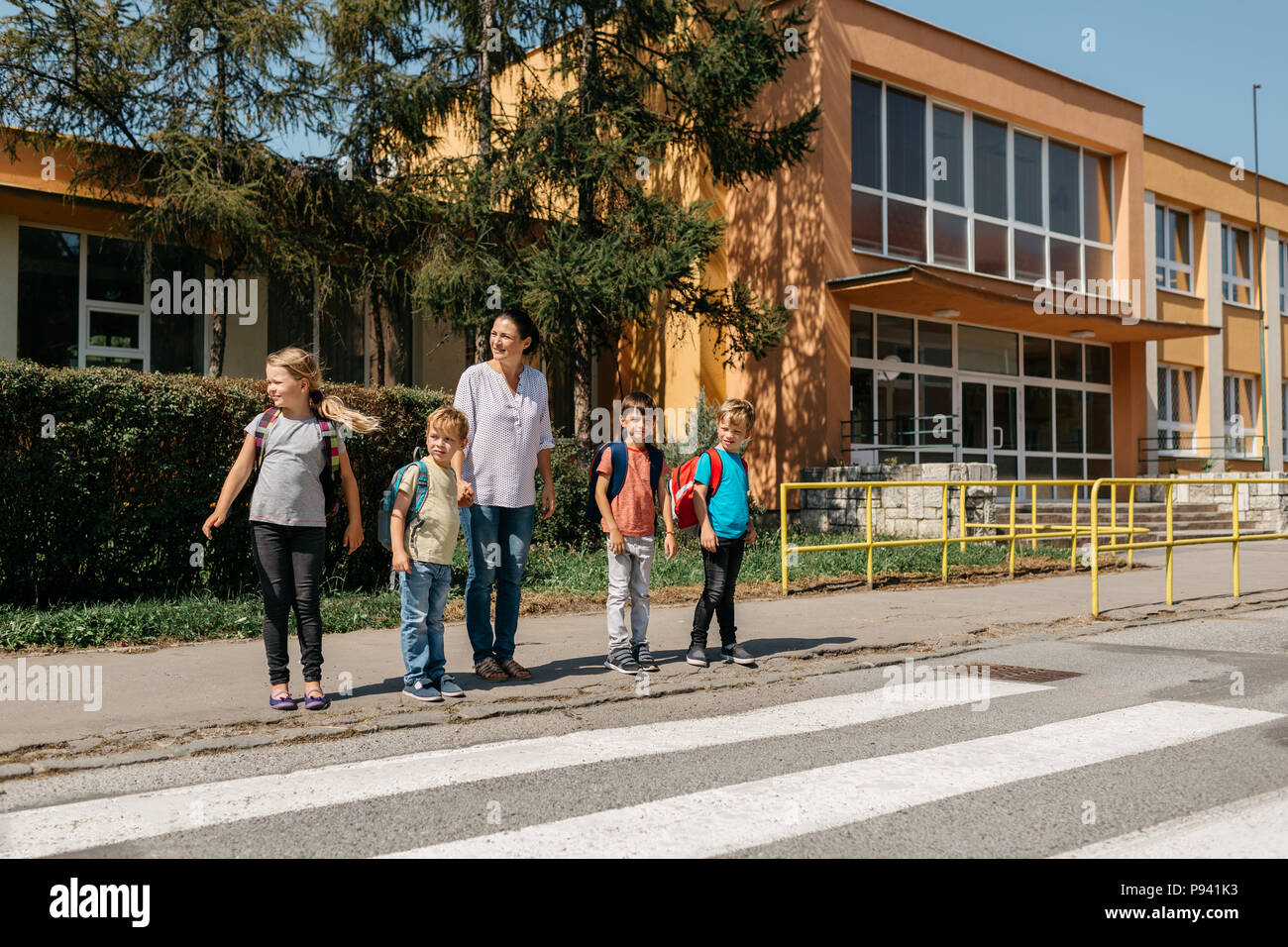 Children waiting at a pedestrian crossing with their mother watching out for cars. Mother teaching children road safety. Stock Photo