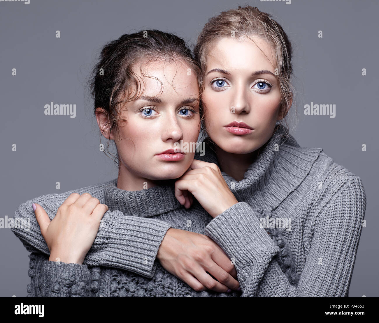 Two young women in gray sweaters on grey studio background. Beautiful girls stretching hands forward in embrace. Female friendship concept. Stock Photo