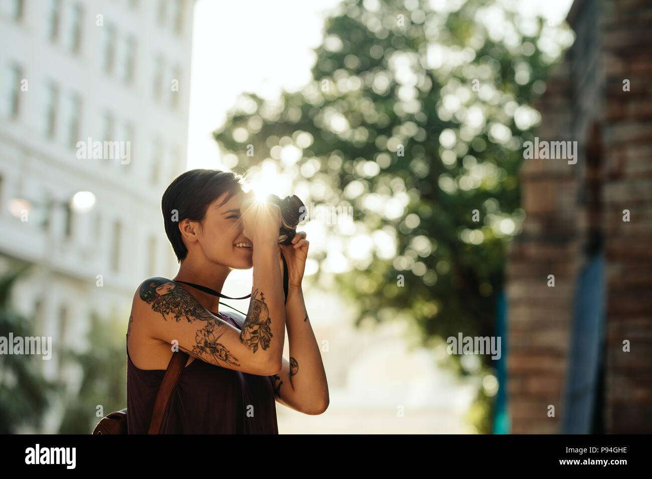 Woman traveler taking photo using her camera with sun in the background. Tourist with tattoo on her hand going around the city taking photographs. Stock Photo