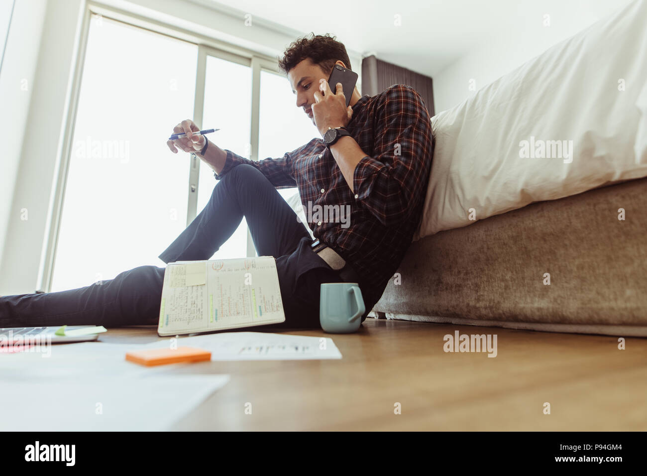 Man working on laptop computer sitting on the floor at home. Freelancer sitting on floor beside a bed and talking over mobile phone looking at his dia Stock Photo
