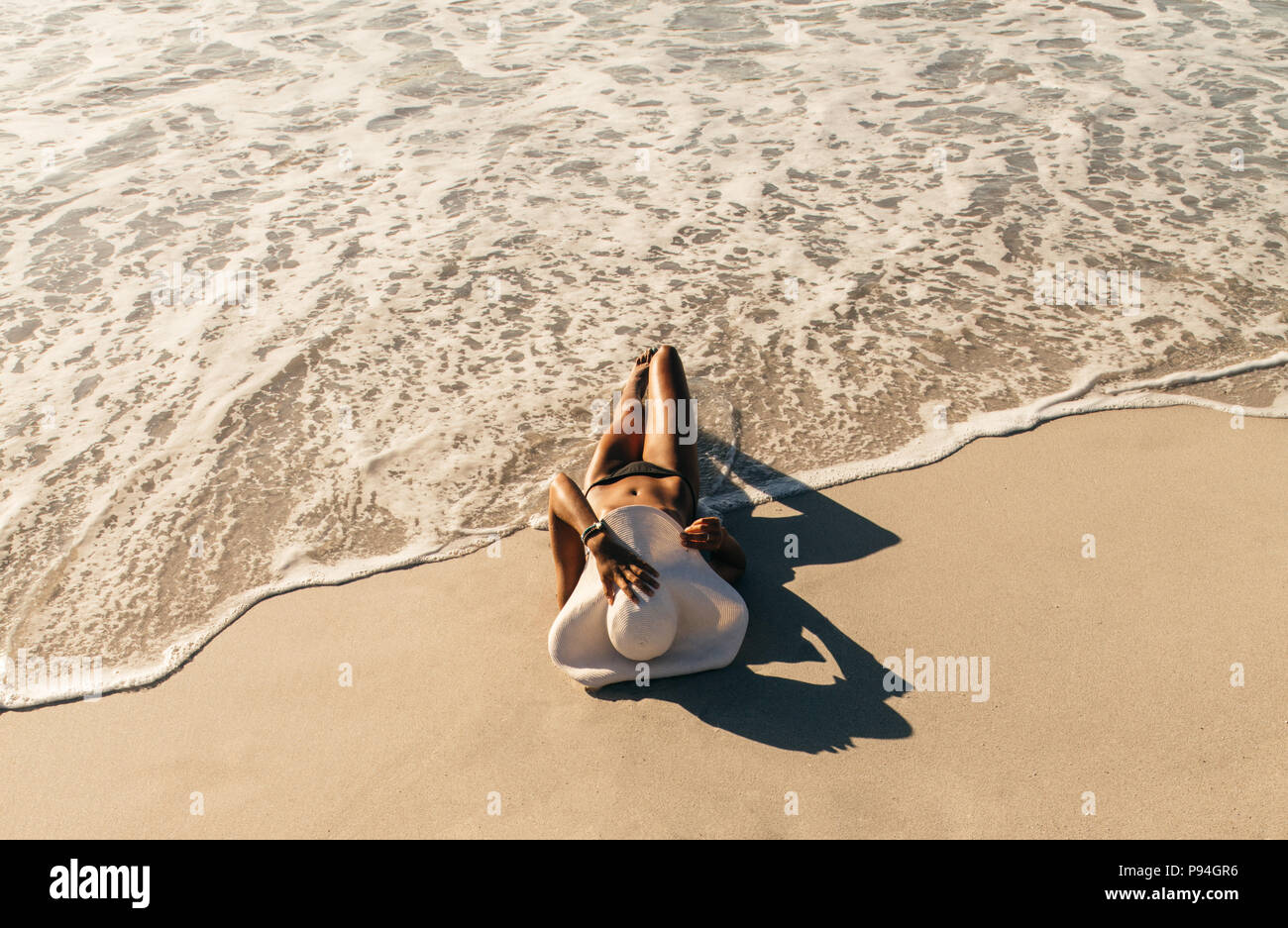 Woman in bikini sunbathing lying near the seashore wearing sun hat with sea waves touching her. Woman on vacation wearing sun hat relaxing at the beac Stock Photo