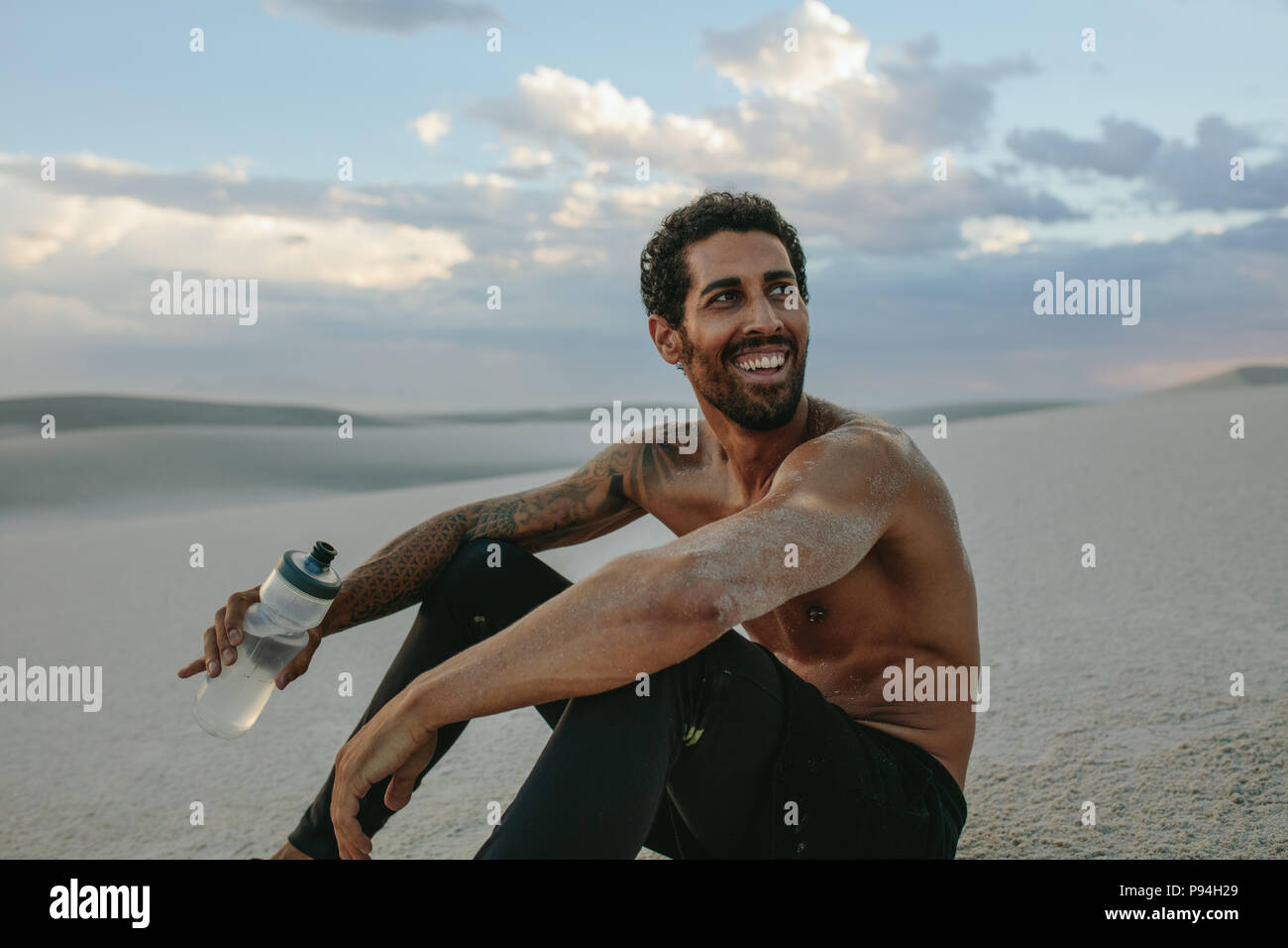 Smiling muscular male athlete sitting on desert sand with a water ball in hand. Tired man taking a break from training in desert. Stock Photo