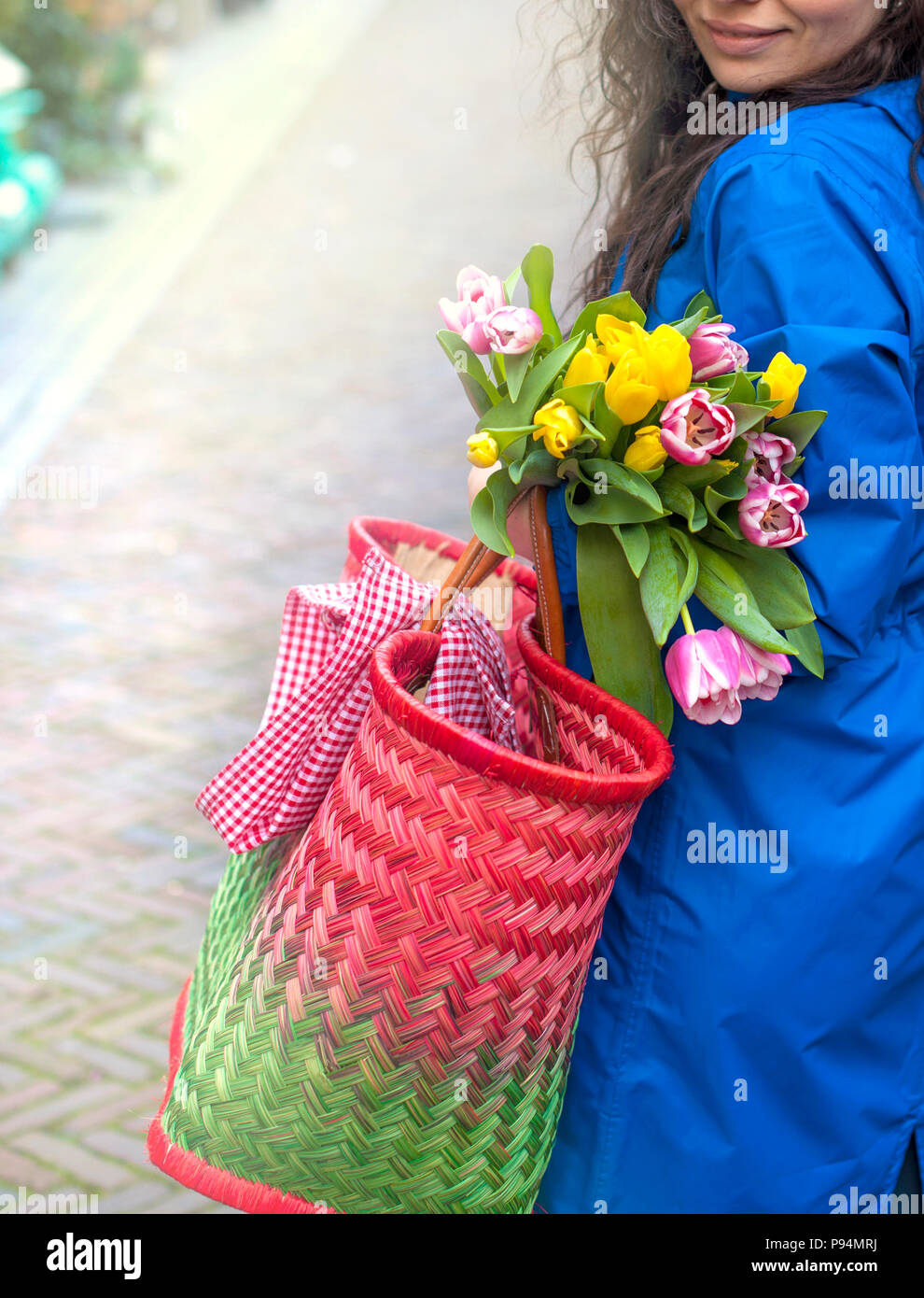girl with a bouquet of spring tulips in pink. In a blue raincoat and yellow boots on the street in Europe. Happy woman. Smile on the face. Card. Stock Photo