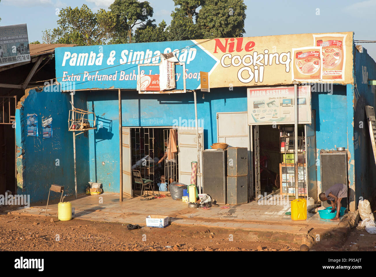 Wakiso Town, Uganda - Street scene. Store shops. A boy is washing clothes in front of a retail store. Stock Photo