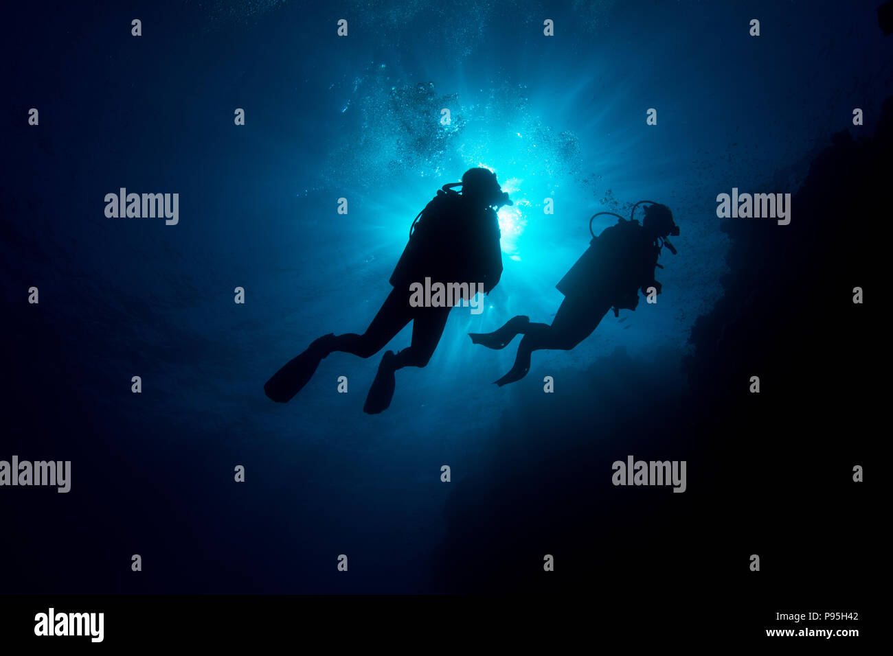 Two scuba divers exploring a tropical coral reef are silhouetted against the surface of the ocean and sunburst in background Stock Photo