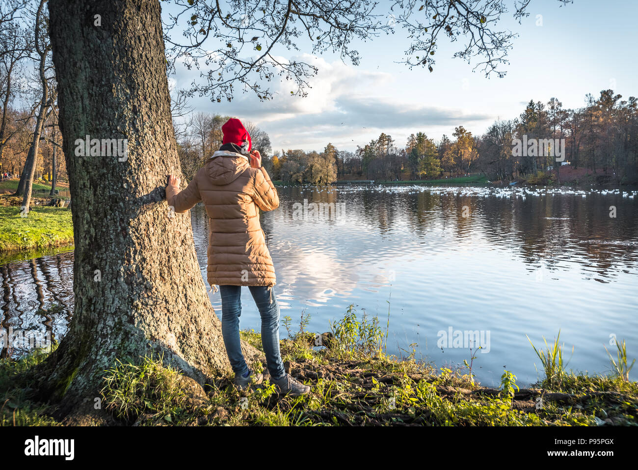 A girl in a red hat wearing headphones enjoys the view of an autumn park. View from the back. Stock Photo