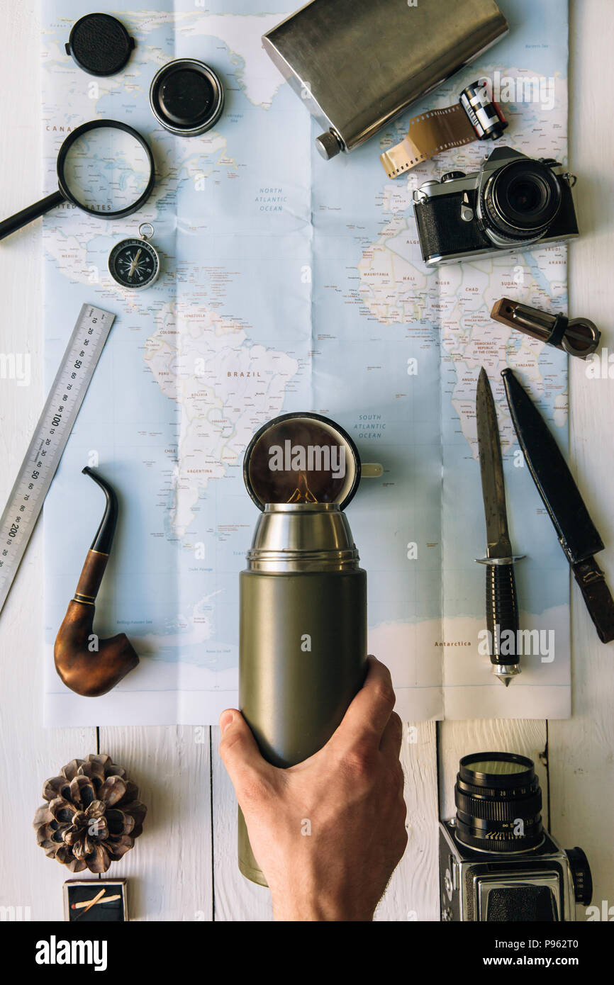 Adventure planning flat lay. Travel vintage gear on map. Traveler, explorer hands in frame pouring coffee or tea from thermos into mug. Vertical explo Stock Photo