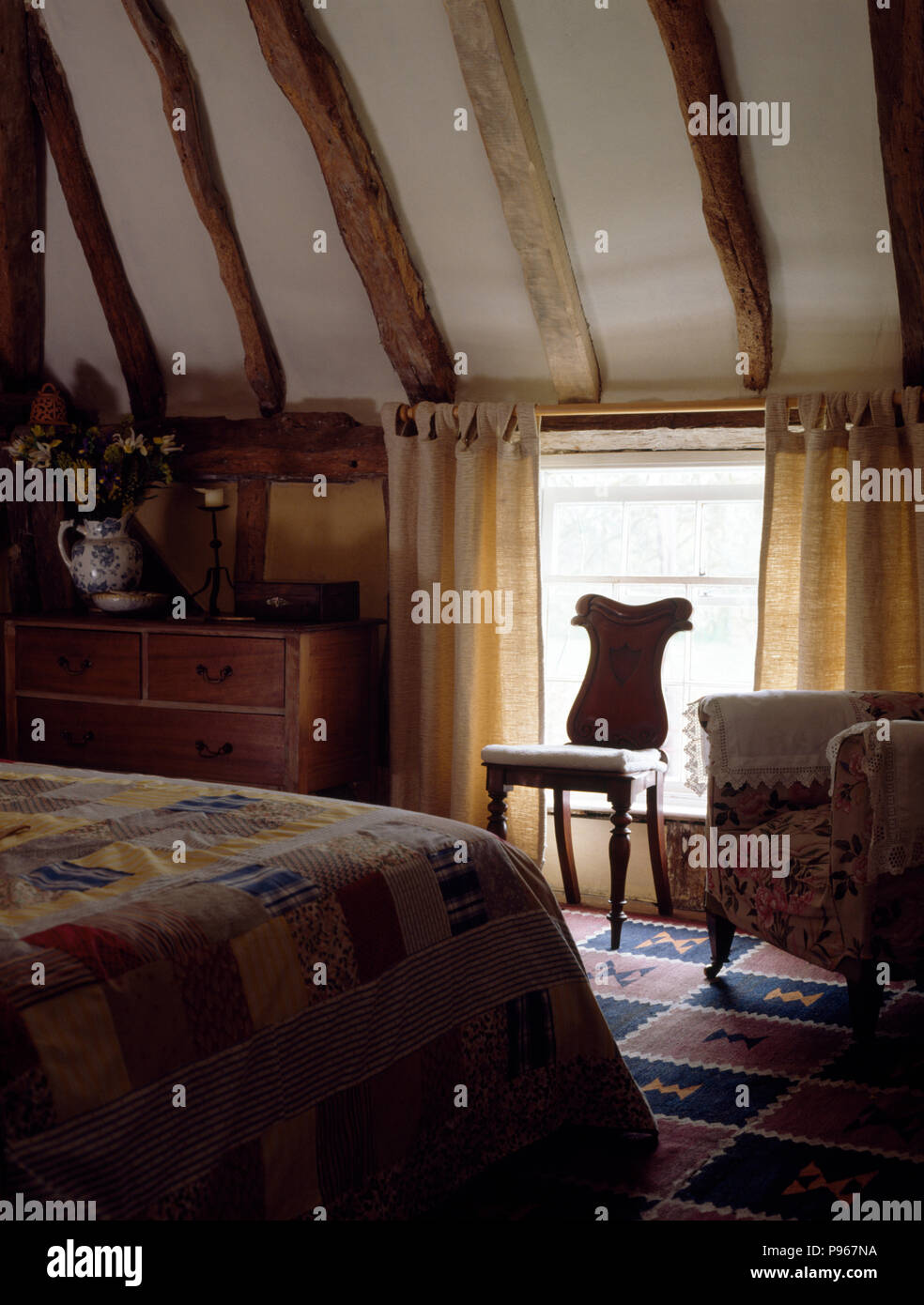Patchwork quilt on bed in attic bedroom with antique chair in front of window with natural linen curtains Stock Photo