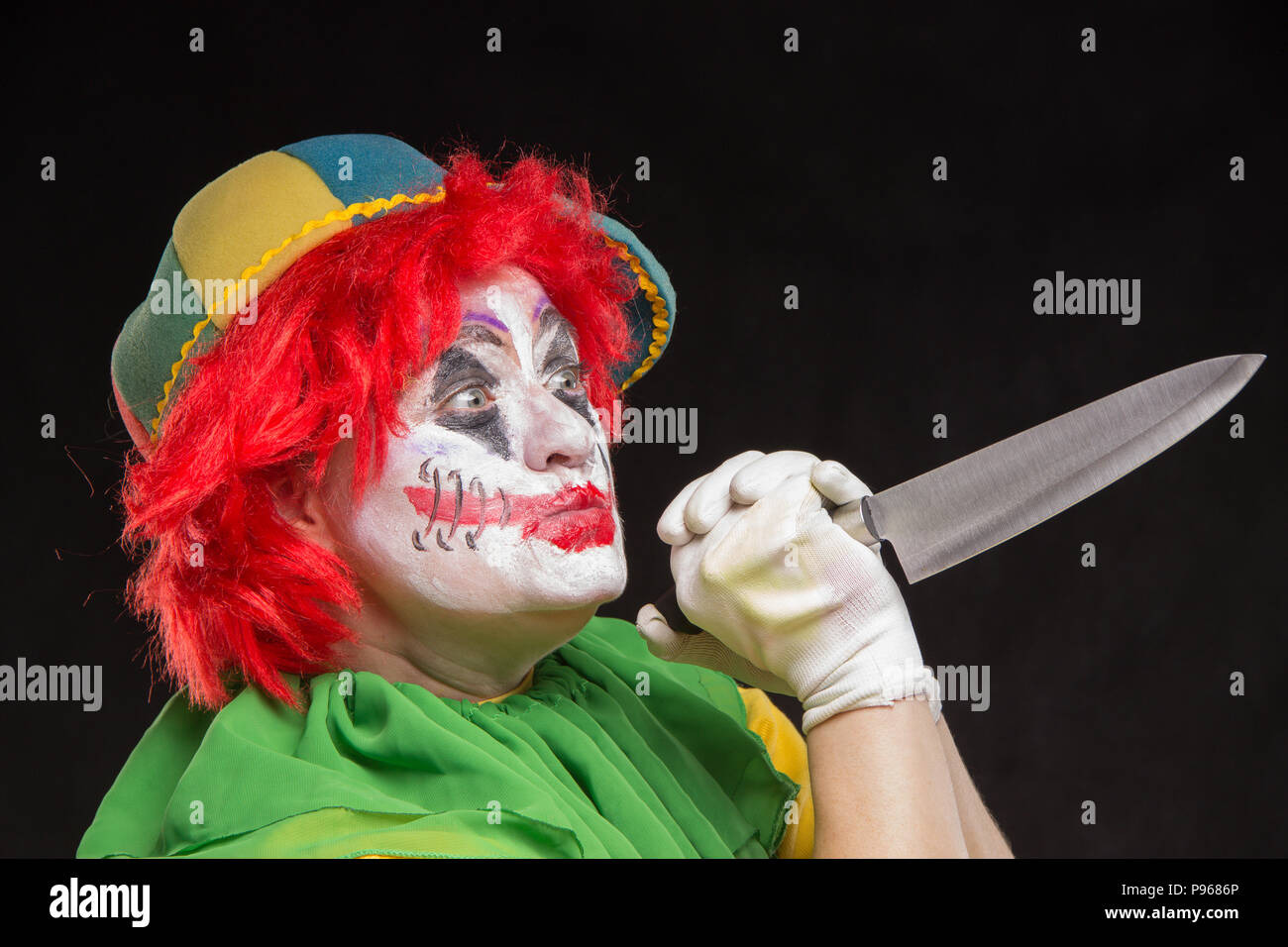Scary clown with a horrible make-up laughs and with big knives in hands on a black background Stock Photo