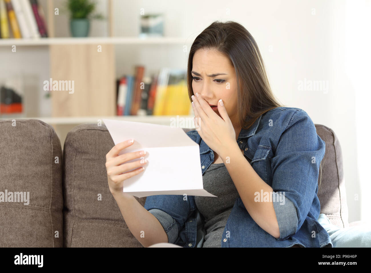 Sad woman reading bad news in a paper letter sitting on a couch in the living room at home Stock Photo